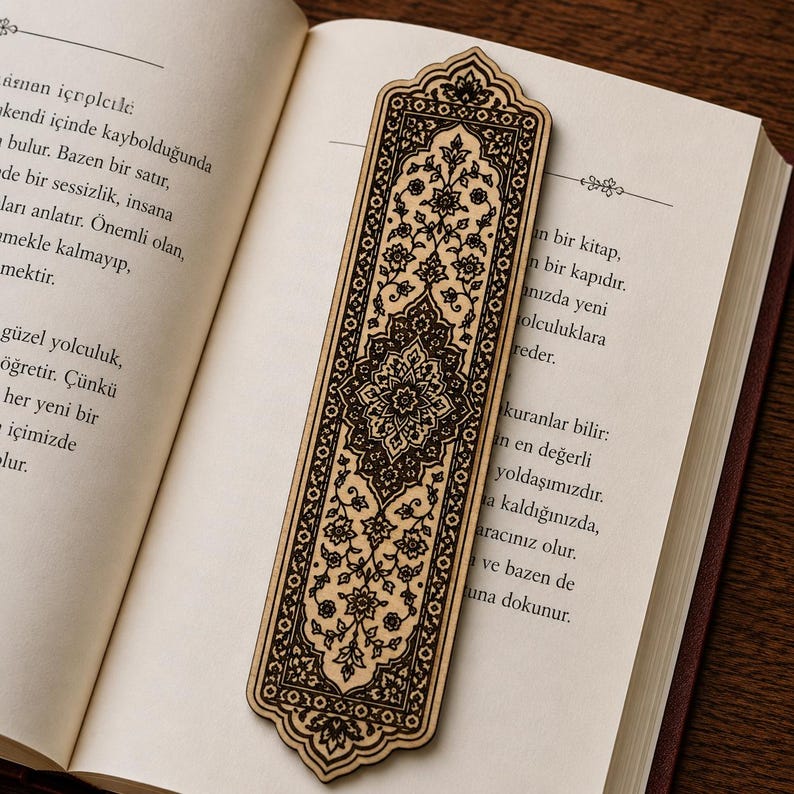 May include: A rectangular wooden bookmark with a pointed arch top and bottom, featuring a dark brown laser-engraved floral and geometric pattern on a light tan wood surface, resting vertically on the open pages of a book with printed text.