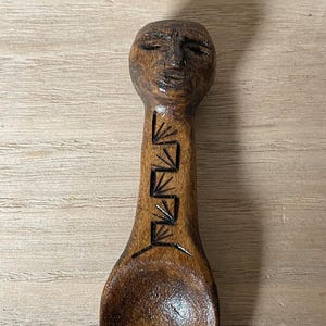 May include: A hand-carved wooden spoon with a face carved into the handle. The spoon is brown with dark brown carved details. The handle features a series of geometric shapes. The spoon's bowl is oval-shaped.