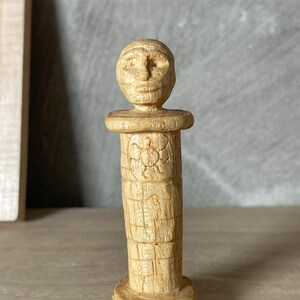 May include: A carved wooden sculpture of a stylized human figure. The sculpture has a cylindrical body with a carved face at the top. A turtle is carved into the body. The sculpture is light brown in color.