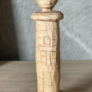 Può includere: Scultura in legno intagliata a mano di una figura con un volto stilizzato in cima a una colonna. La colonna presenta un simbolo ankh intagliato. La scultura è realizzata in legno chiaro e ha un aspetto strutturato a blocchi.
