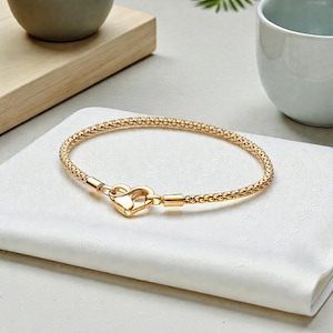 May include: A gold bracelet featuring a heart-shaped clasp. The bracelet has a textured, woven pattern. The clasp is designed as a heart. The bracelet is displayed on a white cloth.