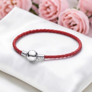 May include: A red braided leather bracelet with a silver clasp. The bracelet is displayed on a white surface, with soft pink roses in the background. The bracelet is a stylish accessory.