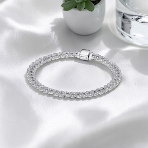 Può includere: Un braccialetto color argento con un design a catena cubana, incastonato con piccoli cristalli scintillanti. Il braccialetto è chiuso con una chiusura rettangolare. Il gioiello è esposto su una superficie bianca, simile al raso, con una piccola pianta in vaso e un bicchiere d'acqua sullo sfondo.