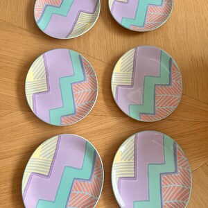 Op de afbeelding: Zes ronde decoratieve borden met een pastelkleurenschema. Elk bord heeft een zigzagpatroon in lichtblauw en lavendel, met gele en koraalaccenten. De borden zijn gerangschikt op een houten oppervlak.