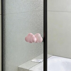 Puede incluir: Dos pomos de armario rosas con forma de nube montados en una puerta de cristal texturizado. Los pomos son de un rosa pastel suave y añaden un toque caprichoso a la decoración del baño. La puerta está enmarcada en negro.