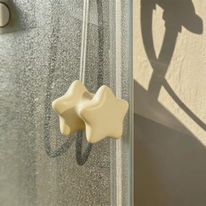 Puede incluir: Dos pomos de armario con forma de estrella, de color crema, están montados en una puerta de ducha de cristal. Los pomos tienen un diseño redondeado y tridimensional. El fondo muestra una superficie de cristal texturizada y una sombra.