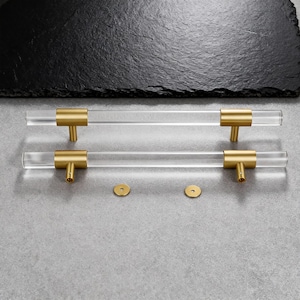 Könnte beinhalten: Zwei transparente Acryl-Schrankgriffe mit goldfarbenen Metallakzenten. Die Griffe sind zylindrisch mit rechteckigen goldfarbenen Endkappen und einer T-förmigen goldfarbenen Basis. Zwei kleine goldfarbene Unterlegscheiben sind ebenfalls vorhanden.