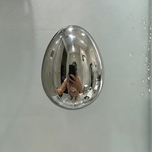 Puede incluir: Un objeto cromado brillante en forma de huevo está montado sobre una superficie de vidrio esmerilado. El objeto refleja el entorno, incluyendo a una persona sosteniendo un teléfono. Gotas de agua son visibles en el vidrio.