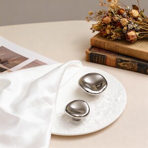May include: Two polished silver cabinet knobs with a smooth, organic shape rest on a white textured plate. The knobs have a reflective surface, and the plate is set against a neutral background. Decorative hardware for home decor.