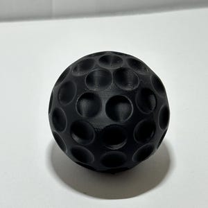 Op de afbeelding: Een zwarte golfbal met een putjesoppervlak. De bal is rond en heeft een matte afwerking. De putjes zijn gelijkmatig verdeeld over het oppervlak van de bal.