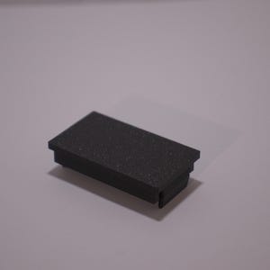 May include: A small, rectangular, black object with a textured surface. The object has a slightly raised edge around the top. The item is likely a component or part of a larger product.