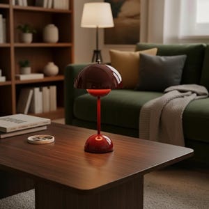 Puede incluir: Una lámpara de mesa roja y granate con una pantalla redondeada y un tallo delgado se encuentra sobre una mesa de centro de madera oscura. La lámpara está iluminada, proyectando un brillo cálido. Un sofá verde y una estantería están al fondo.