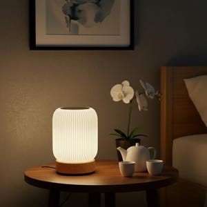 Peut inclure: Une lampe de chevet blanche avec un abat-jour côtelé est posée sur une table ronde en bois. La lampe est associée à une base en bois. Une théière blanche, deux tasses et une orchidée en pot sont également sur la table. Une image encadrée est accrochée au mur.
