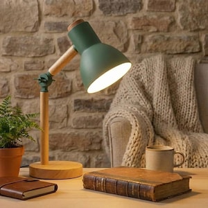 May include: Green and wood desk lamp with a round wooden base. The lamp has a green lampshade and a wooden arm. A fern in a terracotta pot, a leather-bound journal, and a vintage book are on the table.
