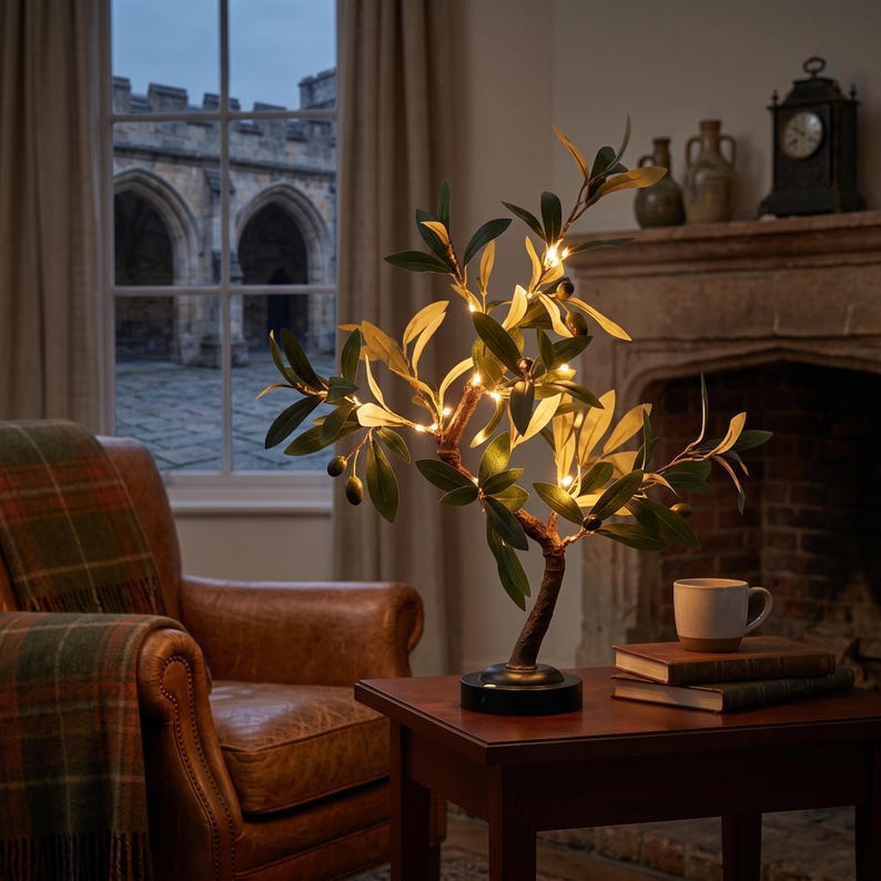 May include: An olive tree-shaped decorative lamp with warm white LED lights. The lamp has green leaves and small olive-like details. It sits on a dark base on a wooden table. The background includes a leather armchair, books, and a fireplace.