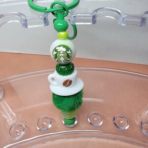 May include: A green and white Starbucks-themed keychain. The keychain features a green ring, a green and white beaded design with the Starbucks logo, a coffee cup, and a green fluffy base. The keychain is designed to be a decorative accessory.