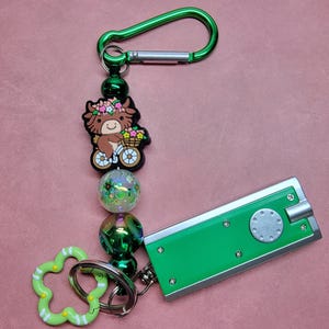 May include: A green keychain with a green carabiner clip, a cow charm riding a bicycle, iridescent and green beads, and a green flower-shaped clip. A green flashlight is attached to the keychain.