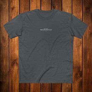 May include: A charcoal gray t-shirt with the text "Error 404: Motivation Not Found" in a small, centered font. The shirt has a crew neck and short sleeves. The background is a wooden surface.