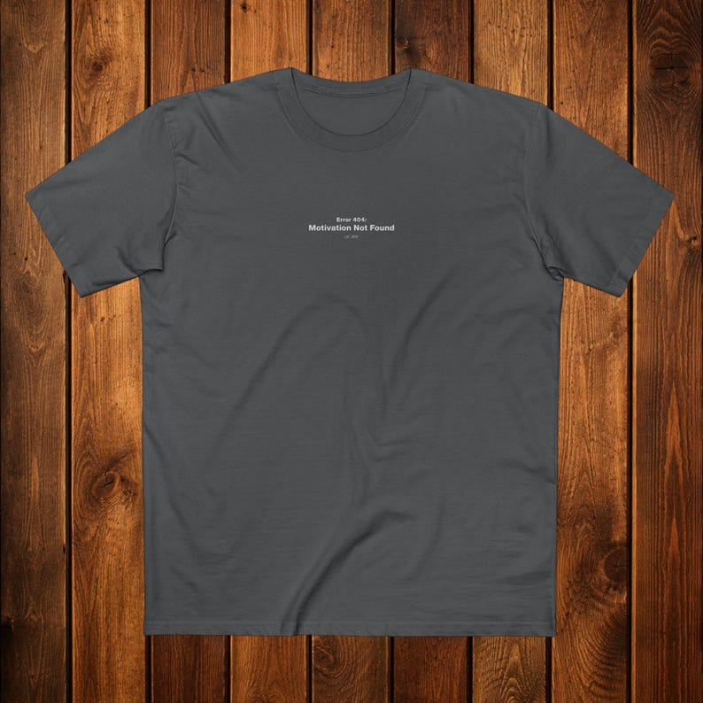 May include: A charcoal gray t-shirt with the text "Error 404: Motivation Not Found" in a small, centered font. The shirt has a crew neck and short sleeves. The background is a wooden surface.