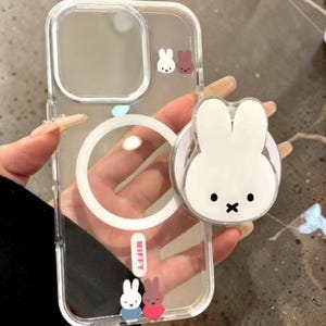 May include: Transparent phone case featuring a white bunny pop socket and small bunny illustrations. The case has a white circle and the word "MIFFY" in a red rectangle. The case is clear.