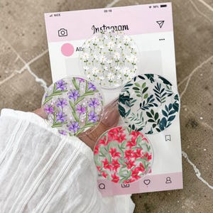 Può includere: Quattro supporti rotondi e trasparenti per telefono con disegni floreali. Uno presenta margherite bianche, un altro iris viola, un terzo foglie verdi e l'ultimo gigli rossi. Su sfondo rosa a tema Instagram.