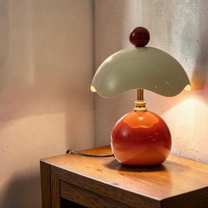 May include: A table lamp featuring a glossy orange spherical base and a pale green, scalloped, dome-shaped shade. A small, dark brown spherical finial sits atop the shade, which is supported by a slender, metallic gold-toned central stem.