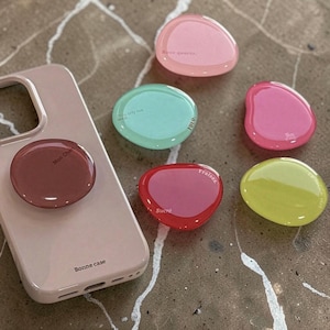 May include: A pale pink phone case with a matching pop socket and the text "Bonne case". Six colourful, pebble-shaped phone grips in pink, green, red, and yellow are scattered around the case. Each grip has a different word.