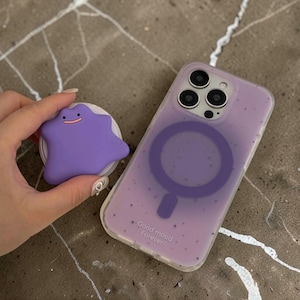 Könnte beinhalten: Eine lilafarbene Handyhülle mit passendem Pop-Socket mit einem Ditto-Design. Die Hülle hat eine transparente Rückseite mit einem lilafarbenen Magnetring und dem Text "Good mood Forever". Der Pop-Socket wird in einer Hand gehalten.