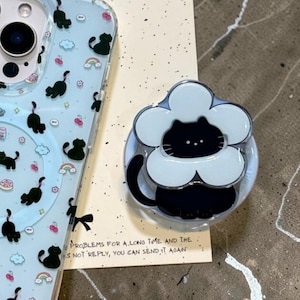 Può includere: Una custodia trasparente per telefono con sfondo azzurro e disegni di gatti neri, arcobaleni e nuvole. È visibile anche un supporto per telefono a forma di fiore abbinato con un design di gatto nero. Il testo sulla carta recita: "PROBLEMS FOR A LONG TIME AND THE AS NOT REPLY, YOU CAN SEND IT AGAIN."