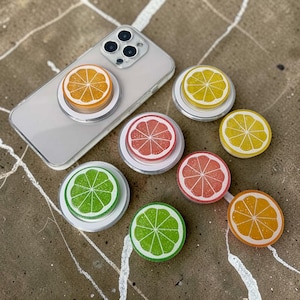 May include: A collection of fruit-themed phone grips and a smartphone. The grips feature orange, lemon, lime, and grapefruit slice designs. The phone has a clear case, and one grip is attached. The grips are white with colorful fruit designs.