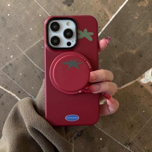 May include: A red phone case with a tomato design. The case features a pop-up grip shaped like a tomato with a green stem, and a small green stem design on the top. The words "Lovely power" are printed on the bottom of the case.