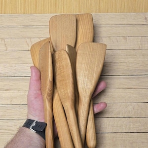 May include: A collection of light brown wooden spatulas with long, smooth handles and flat, rounded blades. The kitchen utensils feature a natural wood grain pattern and are arranged in a fan shape, showcasing their uniform design and smooth texture.