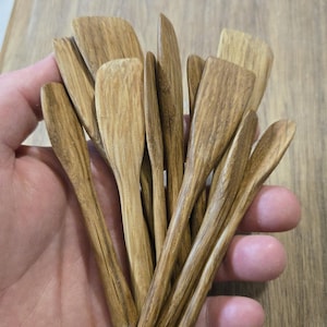 May include: A collection of small, wooden utensils, including spoons and spatulas, held in a hand. The utensils have a natural wood grain and a light brown color. The items appear to be handmade and are likely for kitchen use.