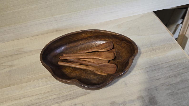 Pode incluir: Uma tigela de madeira castanha escura com uma forma org&acirc;nica, cheia de v&aacute;rias colheres pequenas de madeira. As colheres e a tigela t&ecirc;m um tom quente, sugerindo um acabamento de madeira natural. As colheres t&ecirc;m cerca de 15 cm de comprimento.