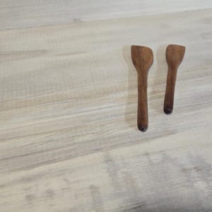 Pode incluir: Duas pequenas colheres de madeira artesanais com acabamento castanho escuro. As colheres t&ecirc;m um design simples e minimalista, com uma cabe&ccedil;a plana e retangular e um cabo longo e fino. S&atilde;o exibidas numa superf&iacute;cie de madeira clara.