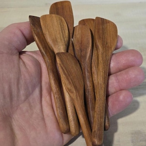 May include: A collection of small, wooden spoons with a natural, warm brown color. The spoons have a smooth finish and a rounded bowl shape, perfect for measuring spices or serving condiments. The spoons are held in a hand.