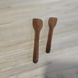Pode incluir: Duas pequenas colheres de madeira artesanais com um acabamento castanho escuro rico. As colheres t&ecirc;m um design simples e elegante, perfeito para servir ou mexer. As colheres s&atilde;o exibidas numa superf&iacute;cie de madeira de cor clara.