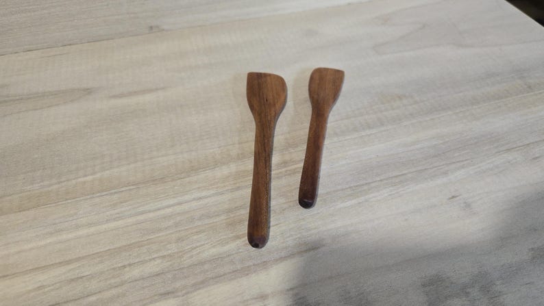 Pode incluir: Duas pequenas colheres de madeira artesanais com um acabamento castanho escuro rico. As colheres t&ecirc;m um design simples e elegante, perfeito para servir ou mexer. As colheres s&atilde;o exibidas numa superf&iacute;cie de madeira de cor clara.