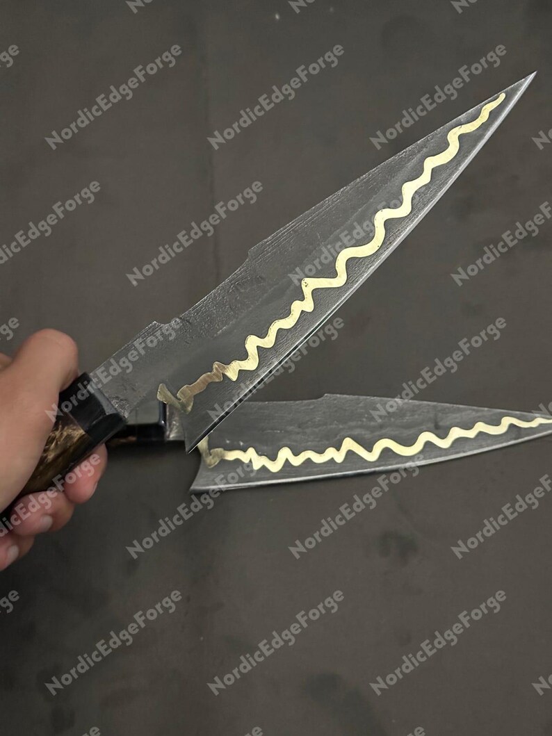 K&ouml;nnte beinhalten: Zwei handgefertigte Messer mit dunklen Klingen und goldfarbenen, wellenf&ouml;rmigen Akzenten. Die Griffe sind dunkelbraun. Die Messer werden vor einem dunklen Hintergrund pr&auml;sentiert. Der Text "Nordic Edge Forge" ist im Hintergrund sichtbar.