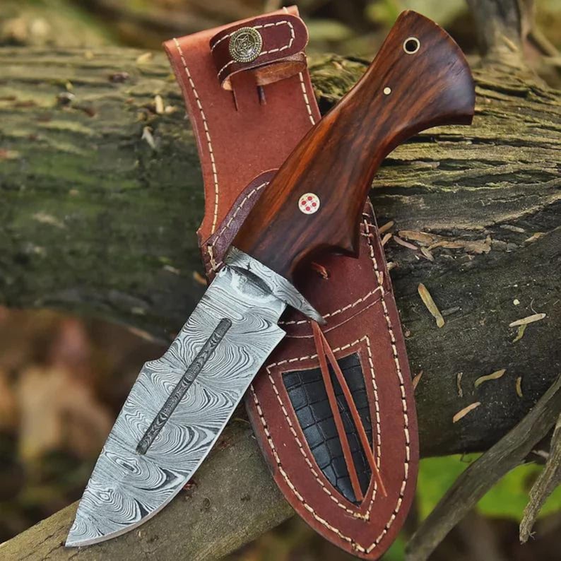 K&ouml;nnte beinhalten: Ein Jagdmesser aus Damaststahl mit einem dunklen Holzgriff und einer braunen Lederscheide. Die Klinge hat ein einzigartiges Wirbelmuster. Die Scheide hat einen dekorativen Knopf und N&auml;hte.