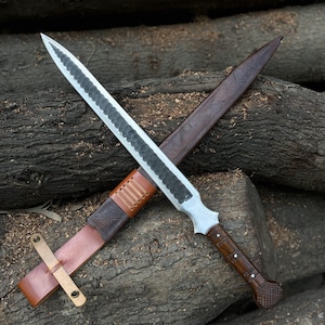 Puede incluir: Una espada artesanal con una hoja plateada y un mango de madera oscura, apoyada sobre una superficie de madera. La espada se combina con una funda de cuero marrón y un cinturón de cuero. La hoja tiene un diseño texturizado.