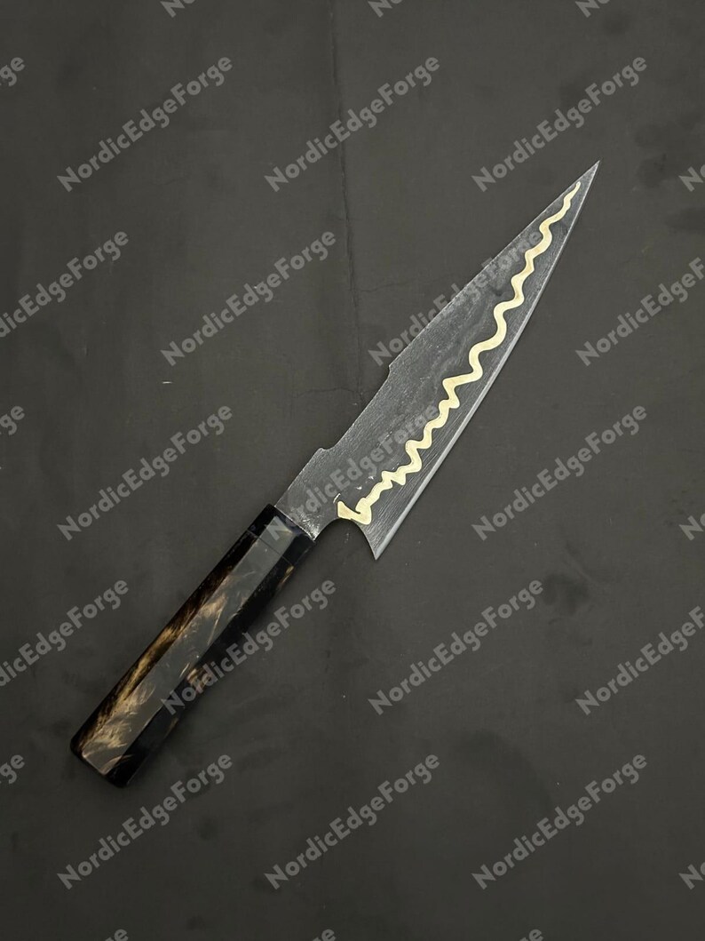 K&ouml;nnte beinhalten: Ein handgefertigtes Messer mit dunkler Klinge und einem dekorativen goldenen Wellenmuster. Der Griff ist schwarz und braun. Der Text "NordicEdgeForge" ist im Hintergrund sichtbar.