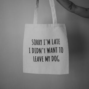 Könnte beinhalten: Eine weiße Stofftasche mit zwei Tragegriffen, die auf der Vorderseite mittig mit dem schwarzen Text "SORRY I'M LATE I DIDN'T WANT TO LEAVE MY DOG" bedruckt ist. Die Tasche besteht aus hellem Stoff und hat ein schlichtes, rechteckiges Design.