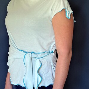 May include: Light blue top with a boat neck and short sleeves. The top features a wrap-around design with a tie at the waist. The edges of the sleeves and the wrap-around tie are trimmed with a contrasting bright blue color.