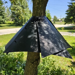 May include: A black, conical tree squirrel baffle made of rigid material, featuring a wrap-around design with overlapping edges, secured by black plastic fasteners and clips, mounted around the trunk of a tree to prevent climbing animals.