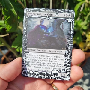 Può includere: Una carta collezionabile rettangolare intitolata Syr Konrad, the Grim, con l'illustrazione di un cavaliere umano, un bordo decorato con teschi in bianco e nero e testo stampato che descrive abilità, forza e costituzione di 5/4.