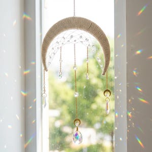 May include: A crescent moon suncatcher with a beige woven frame and hanging crystals. The crystals are suspended from gold chains of varying lengths. The suncatcher hangs in front of a window, with light refracting through the crystals, creating rainbows.