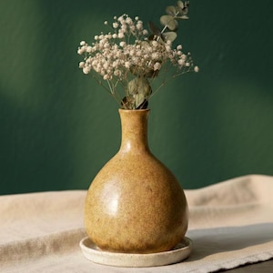 Puede incluir: Un pequeño jarrón de cerámica amarillo-marrón moteado con cuello estrecho y cuerpo redondeado, que contiene un ramo de flores blancas y follaje verde. El jarrón está sobre un plato pequeño de cerámica blanquecina. El fondo es verde oscuro liso.