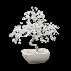 May include: A crystal tree ornament with a twisted silver wire trunk and branches. The tree has clear crystal accents and is in a white square ceramic pot filled with small white stones. The background is black.