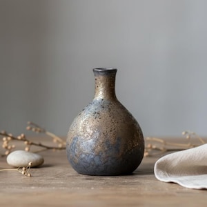 Könnte beinhalten: Eine kleine, handgefertigte Keramikvase mit einer gesprenkelten metallisch goldfarbenen und dunkelblauen Glasur. Die Vase hat einen abgerundeten Körper und einen schmalen Hals. Die Vase wird auf einer Holzoberfläche mit neutralem Hintergrund präsentiert.